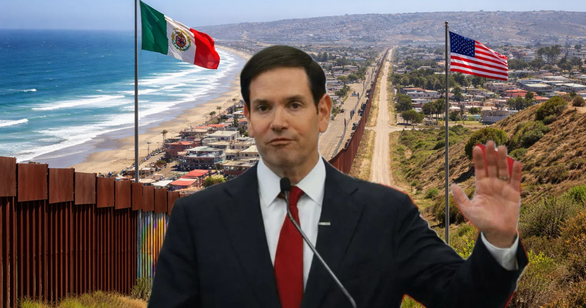 Hombre con micrófono en una rueda de prensa en la frontera México-EE.UU., con banderas de ambos países y una cerca fronteriza de fondo.